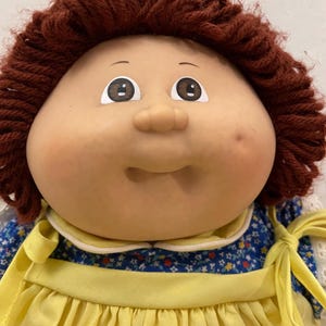 Puede incluir: Una muñeca vintage con una cara redonda, ojos marrones y cabello de hilo castaño rojizo. La muñeca viste un vestido amarillo con un corpiño azul con estampado floral y un cuello blanco. La cara de la muñeca tiene una tez rosada y un pequeño hoyuelo.