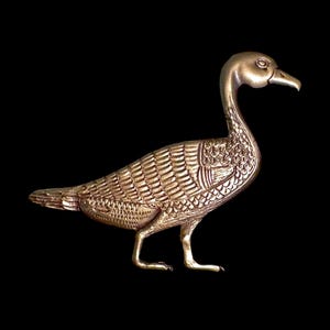 May include: A detailed, golden-colored metal sculpture of a duck or goose. The bird is standing with its head held high, and its feathers are intricately carved. The sculpture is set against a black background.