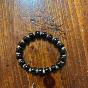 Pode incluir: Um bracelete de contas pretas e douradas. O bracelete é feito de pequenas contas redondas que são enfiadas em um cordão elástico.