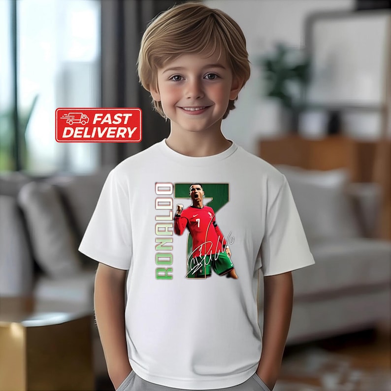 Puede incluir: Camiseta blanca con un gr&aacute;fico de un futbolista con camiseta roja, n&uacute;mero 7. El nombre del jugador, "Ronaldo", est&aacute; impreso verticalmente en verde. La camiseta tiene cuello redondo y mangas cortas.