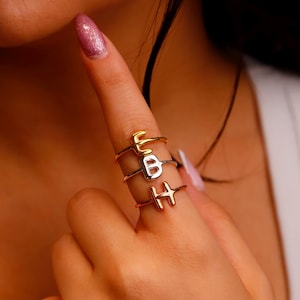 May include: Three delicate rings spelling out the letters "EBH" are stacked on a finger. The rings are in gold, silver, and rose gold tones. The background is blurred, focusing on the jewelry.