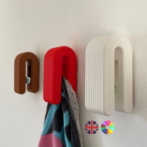 May include: Three U-shaped wall hooks in brown, red, and white. The red hook holds a colourful towel. The hooks are mounted on a white wall, offering a modern storage solution for bathrooms or kitchens.