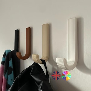 May include: Four J-shaped wall hooks in black, brown, beige, and white, mounted on a white wall. The hooks are holding a scarf and a black bag. The hooks are made of a ribbed material. Decorative stickers are at the bottom.