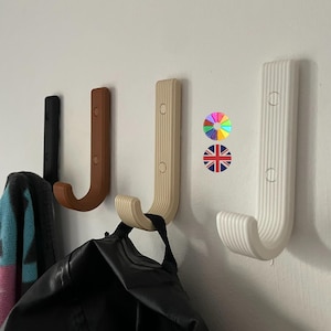 Puede incluir: Cuatro perchas de pared en forma de J en negro, marrón, beige y blanco. Las perchas están montadas en una pared blanca. Una bolsa negra y una toalla colorida cuelgan de dos de las perchas. Las perchas son de material acanalado.