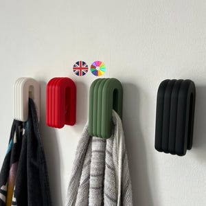 May include: Four wall-mounted towel hooks in white, red, green, and black. Each hook has a U-shaped design with vertical ridges. Towels hang from the hooks. The hooks are mounted on a white wall.
