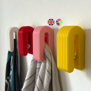 May include: Four U-shaped wall hooks in white, red, pink, and yellow. Each hook has a ribbed design and is mounted on a white wall. Scarves and a towel hang from the hooks, showcasing their functionality.
