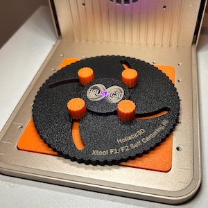 May include: A black circular self-centering jig with four orange adjustable knobs, featuring the text "Holistic3D" and "Xtool F1/F2 Self Centering Jig" printed on the surface, positioned on a metallic base with an integrated ruler.