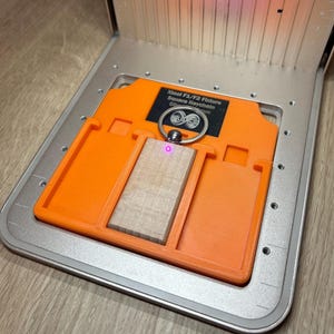 Può includere: Un portachiavi rettangolare in legno chiaro con un anello in metallo argentato, posizionato all'interno di un supporto in plastica arancione con l'etichetta 'Xtool F1/F2 Fixture Square Keychain'. Il supporto è su una base in metallo argentato con ventola.