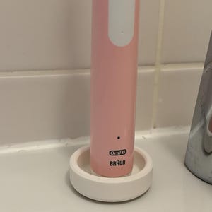 Peut inclure: Un manche de brosse à dents électrique rose clair avec un bouton d'alimentation blanc et une base de chargement circulaire blanche. Les logos Oral-B et Braun sont imprimés en noir sur le bas du manche, positionné verticalement sur son socle.