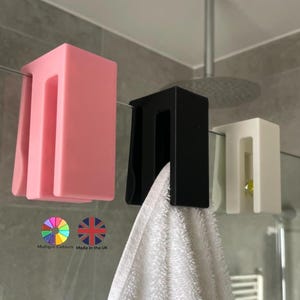 May include: Three towel holders in pink, black, and white are mounted on a glass shower door. A white towel hangs from the black holder. The image includes text indicating multiple colors and that the product is made in the UK.