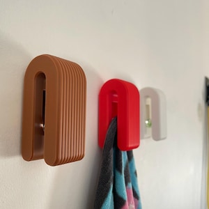 May include: Three U-shaped wall hooks in terracotta, red, and white colours. Each hook features a ribbed, layered design. A grey, pink, and blue patterned towel hangs from the red hook, while the white hook contains a small green bead.