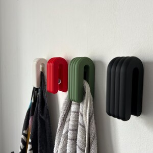 May include: Four U-shaped towel hooks in white, red, green, and black are mounted on a white wall. Each hook has a ribbed design and holds a towel or cloth. The hooks are a modern, minimalist design.