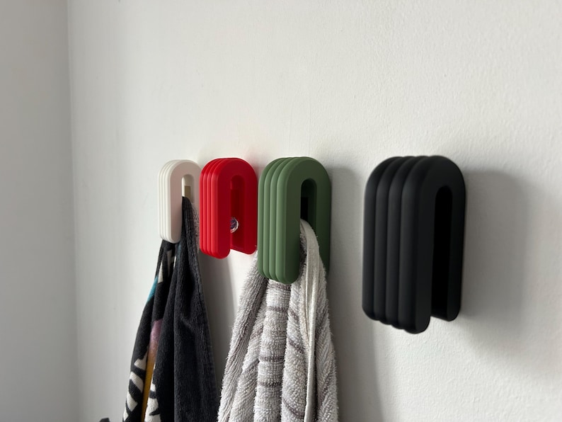May include: Four U-shaped towel hooks in white, red, green, and black are mounted on a white wall. Each hook has a ribbed design and holds a towel or cloth. The hooks are a modern, minimalist design.