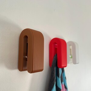May include: Three arch-shaped wall hooks in brown, red, and white. Each hook features a ribbed, layered design. A patterned fabric item hangs from the red hook, while a small green circular accent is visible on the white hook.