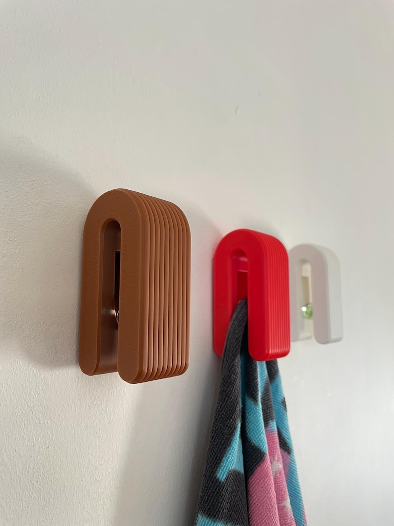 May include: Three arch-shaped wall hooks in brown, red, and white. Each hook features a ribbed, layered design. A patterned fabric item hangs from the red hook, while a small green circular accent is visible on the white hook.