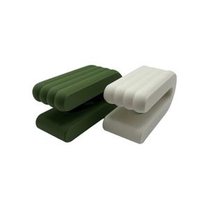 May include: Two modern, minimalist clips in the image. One is a deep green, the other is white. Each clip has a rectangular base and a curved, ridged top. The clips are designed to hold items.