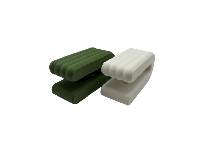 May include: Two modern, minimalist clips in the image. One is a deep green, the other is white. Each clip has a rectangular base and a curved, ridged top. The clips are designed to hold items.