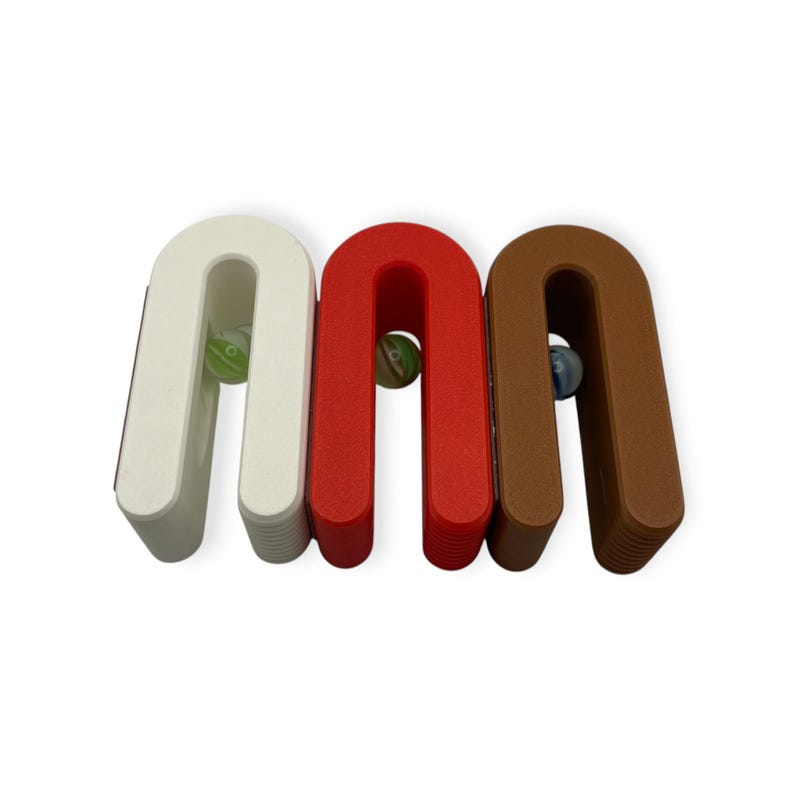 May include: Three U-shaped stands in white, red, and brown, each featuring a textured base and a smooth top arch. A single small glass marble is positioned within the open centre of each stand, showcasing green, white, and blue swirl patterns.