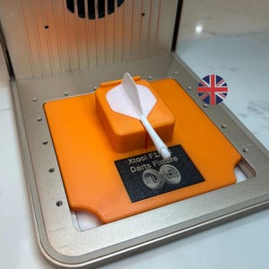 Könnte beinhalten: Eine orangefarbene und silberne Dartvorrichtung mit einem weißen Dart und einem kleinen schwarzen Etikett mit der Aufschrift "Xtool F1 Darts Fixture". Die Vorrichtung dient dazu, einen Dartpfeil an Ort und Stelle zu halten. Der Hintergrund ist hell.