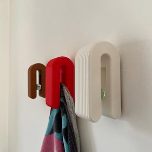 May include: Three U-shaped wall hooks in brown, red, and white, each featuring a ribbed texture on the outer surface. The red hook holds a folded towel with pink, blue, and black patterns. Each hook is secured to the wall with a visible screw.