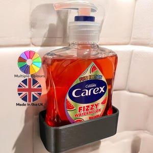 Pode incluir: Um suporte de dispensador de sabonete de plástico preto com uma garrafa de sabonete para as mãos Carex Fizzy Watermelon. A garrafa de sabonete é vermelha e transparente com um rótulo azul e branco. O rótulo diz "Fun Edition" e "Cussons Carex Fizzy Watermelon".