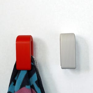 May include: Two U-shaped wall hooks with a vertical ribbed texture. One hook is bright red and holds a patterned towel with blue, black, and pink colours. The other hook is light grey and is empty. Both hooks are mounted on a plain white surface.