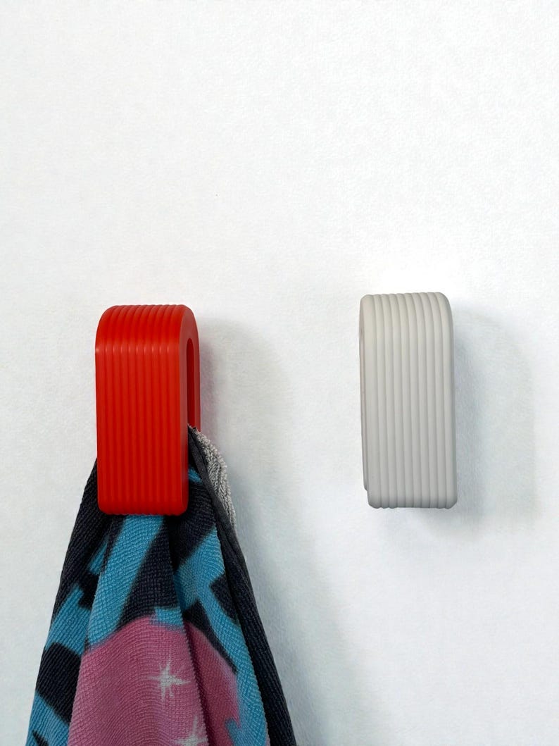 May include: Two U-shaped wall hooks with a vertical ribbed texture. One hook is bright red and holds a patterned towel with blue, black, and pink colours. The other hook is light grey and is empty. Both hooks are mounted on a plain white surface.
