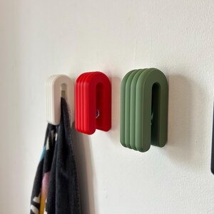 May include: A collection of U-shaped wall hooks in white, red, green, and black. The hooks are mounted on a white wall, with a patterned towel hanging from the white hook. The hooks have a ribbed design.