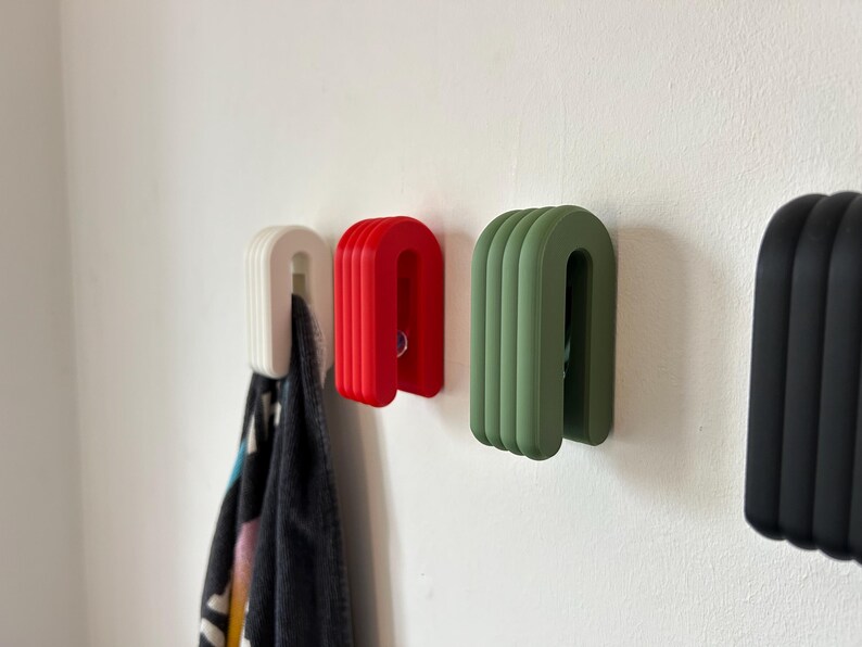 May include: A collection of U-shaped wall hooks in white, red, green, and black. The hooks are mounted on a white wall, with a patterned towel hanging from the white hook. The hooks have a ribbed design.