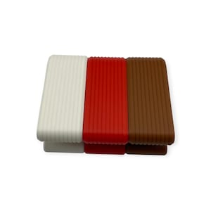 May include: Three rectangular door stops arranged side by side. Each door stop features a ribbed, textured surface and a wedge shape. The colours of the door stops are white, bright red, and dark brown, all made of a solid material.