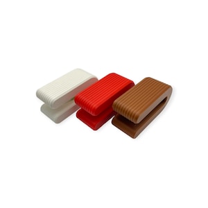 May include: Three U-shaped clips with a ribbed texture, arranged in a row. The clips are coloured white, bright red, and brown. Each clip features a rounded rectangular profile with parallel grooves running along the outer surface.