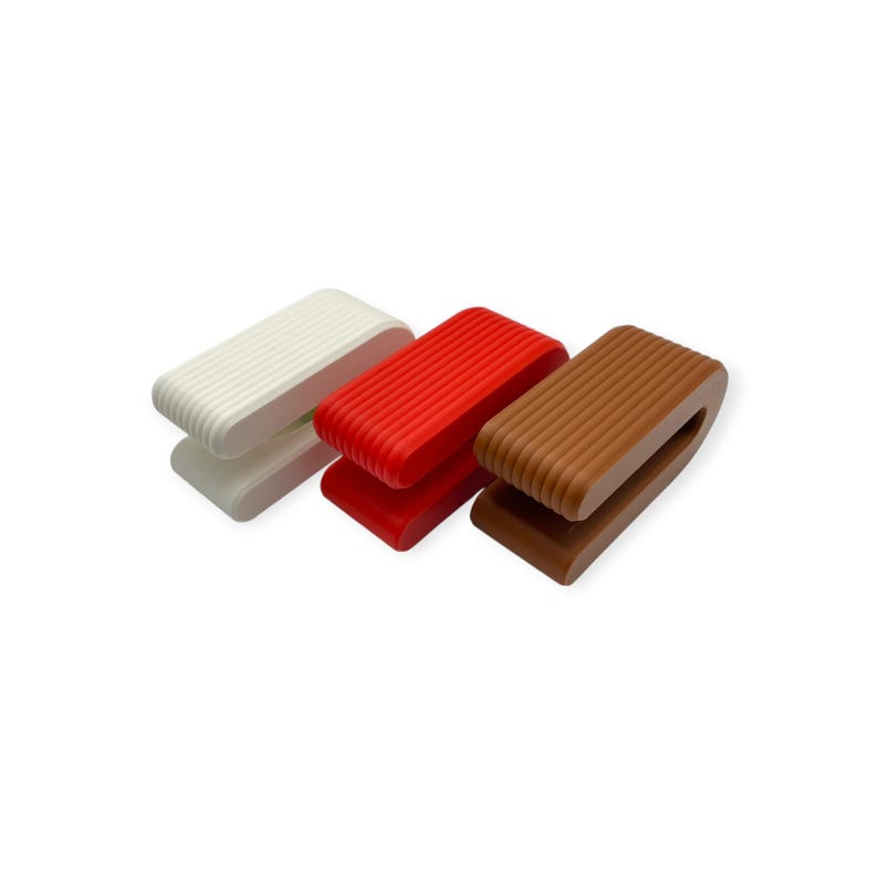 May include: Three U-shaped clips with a ribbed texture, arranged in a row. The clips are coloured white, bright red, and brown. Each clip features a rounded rectangular profile with parallel grooves running along the outer surface.