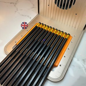 Puede incluir: Un conjunto de lápices negros con férulas doradas y puntas sin afilar. Los lápices están dispuestos sobre una superficie naranja dentro de un marco de metal plateado. Una herramienta de escritura o dibujo.