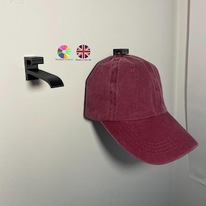 Può includere: Un berretto da baseball bordeaux è appeso a un attaccapanni nero a parete. Il berretto ha una visiera curva e un cinturino in tessuto. Il supporto è in metallo nero. L'immagine include il testo "Multiple Colours" e "Made in the UK".