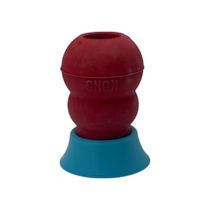 Puede incluir: Un juguete para perros de goma roja con la marca "KONG" grabada en el lateral. El juguete tiene forma bulbosa con una base azul. Este juguete duradero está diseñado para masticar y jugar.