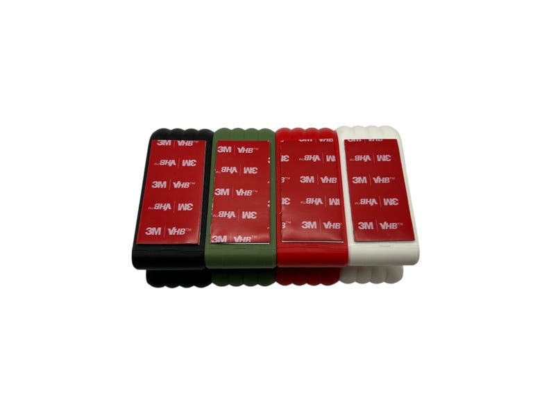 May include: Four rectangular adhesive holders in black, olive green, red, and white. Each has a red adhesive strip with the text "3M VHB". The holders are arranged side by side against a white background.