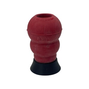 Puede incluir: Un juguete para perros de goma roja con forma bulbosa y base negra. El juguete tiene la marca "KONG" grabada en el lateral. La parte superior tiene una abertura. Este juguete duradero está diseñado para que los perros mastiquen y jueguen.