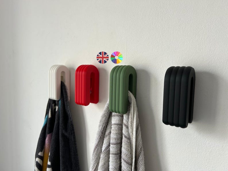 May include: Four wall-mounted towel hooks in white, red, green, and black. Each hook has a U-shaped design with vertical ridges. Towels hang from the hooks. The hooks are mounted on a white wall.