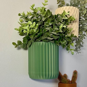 Może przedstawiać: Trzy donice ścienne z zielenią. Zielona, cylindryczna donica z pionowymi grzbietami zawiera bujne zielone liście. Widoczna jest również kremowa donica z rombowym wzorem i mała brązowa donica z kaktusem.