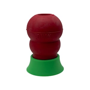 Puede incluir: Un juguete para perros de goma roja con la palabra "KONG" en relieve en el lateral. El juguete tiene una parte superior bulbosa y una bombilla más pequeña debajo, que se asienta sobre una base verde. El juguete está diseñado para perros.