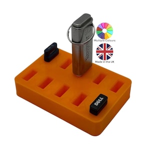 Puede incluir: Un soporte naranja para unidades USB con múltiples ranuras, que contiene una unidad USB Kingston plateada. La imagen también muestra una unidad USB Dell negra y una unidad USB negra. También se ve el texto "Multiple Colours" y "Made in the UK".