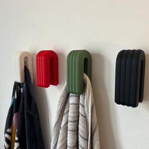 May include: Four modern, U-shaped wall hooks in white, red, olive green, and black. The hooks are mounted on a white wall, with towels hanging from the white and olive green hooks. The hooks have a ribbed design.