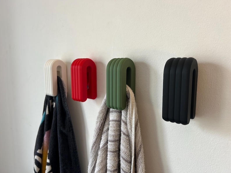 May include: Four modern, U-shaped wall hooks in white, red, olive green, and black. The hooks are mounted on a white wall, with towels hanging from the white and olive green hooks. The hooks have a ribbed design.