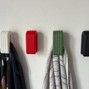 May include: Four modern wall hooks in white, red, green, and black. Each hook has a curved, ribbed design and is mounted on a white wall. Two hooks hold towels, showcasing their functionality and style.