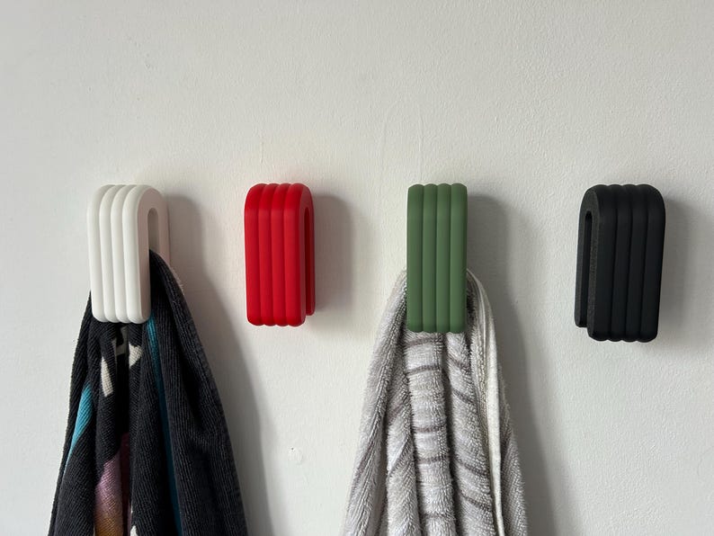 May include: Four modern wall hooks in white, red, green, and black. Each hook has a curved, ribbed design and is mounted on a white wall. Two hooks hold towels, showcasing their functionality and style.