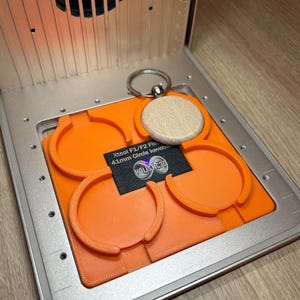 Puede incluir: Una plantilla naranja brillante impresa en 3D con cuatro recortes circulares para llaveros de 41 mm, colocada dentro de una máquina de grabado láser de metal plateado. Un llavero circular de madera clara con una anilla metálica descansa en una ranura.