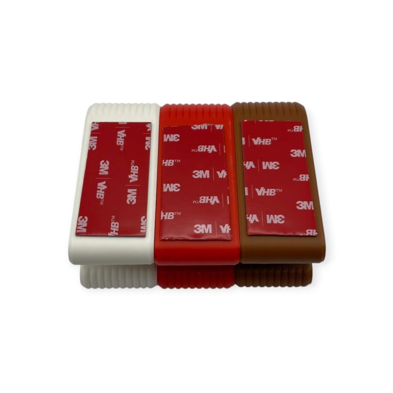 May include: Three rectangular edge guards in white, red, and brown, each featuring a vertical red adhesive strip with the white text 3M VHB printed repeatedly. The guards have a ribbed texture along the sides and a smooth top surface.