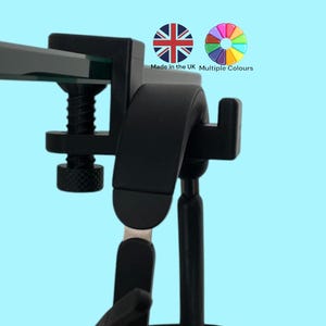 May include: Black clamp-on headset holder with a hook for hanging headphones. The clamp is adjustable and can be attached to a desk or table. The holder is made in the UK and is available in multiple colours.