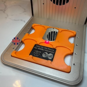 Puede incluir: Un accesorio naranja Xtool F1/F2 para llaveros militares, colocado dentro de un marco de metal plateado. El accesorio tiene el texto "ME005 1.97" x 1.10". Se ilumina una pequeña luz roja.