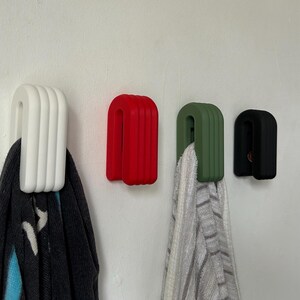 May include: Four wall hooks in white, red, green, and black. Each hook has a rounded, ribbed design. A dark scarf hangs from the white hook, and a striped towel hangs from the green hook. The hooks are mounted on a white wall.
